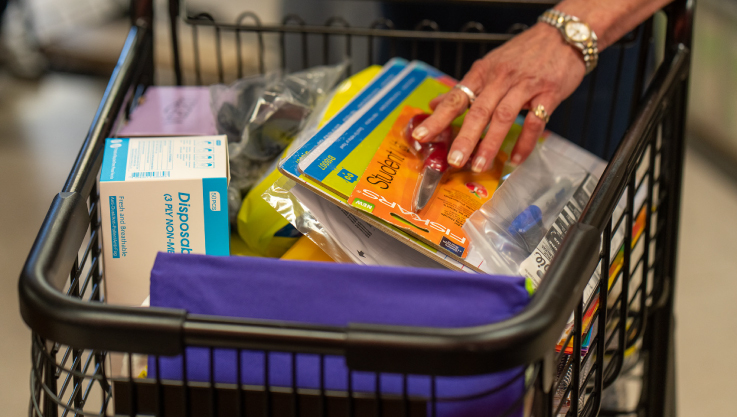 A hand places a pair of scissors in a shopping cart with other school supplies. 