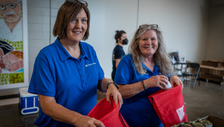 Two women in blue shirts hold red bags filled with food and smile at the camera.