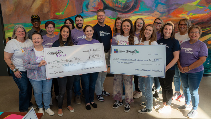 A group of volunteers stand together facing the camera holding two large promotional checks. 