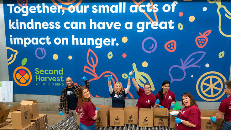 Seven volunteers pack boxes of food on an assembly line. A mural behind them reads "Together, our small acts of kindness can have a great impact on hunger."