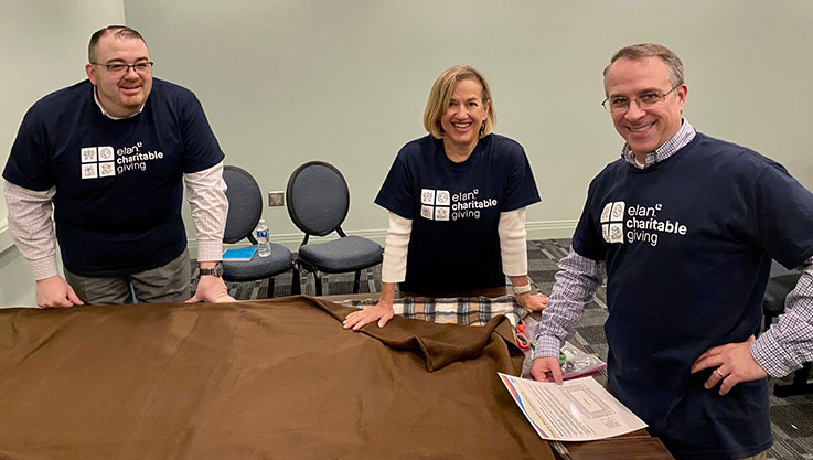 Three adults stand around a table smiling at the camera. On the table are fabric and scissors. 