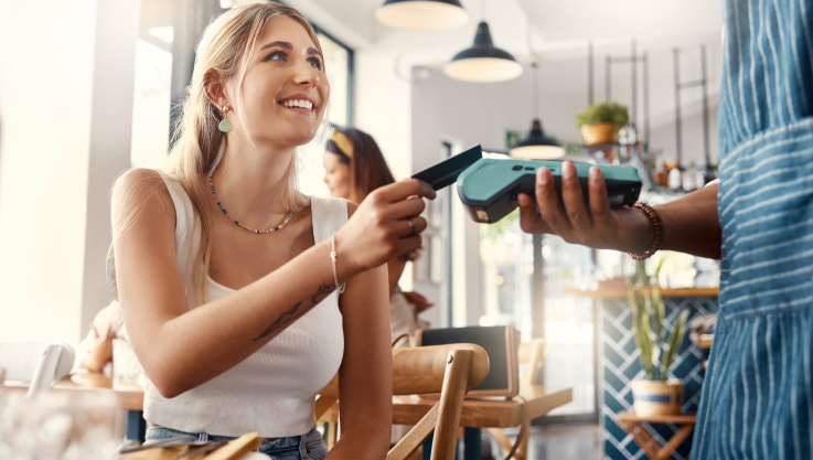 A woman sits at a restaurant table and holds her credit card over a tableside terminal held by the server.