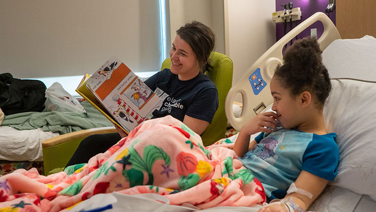 An adult volunteer reads a book at the bedside of a child in a hospital. 