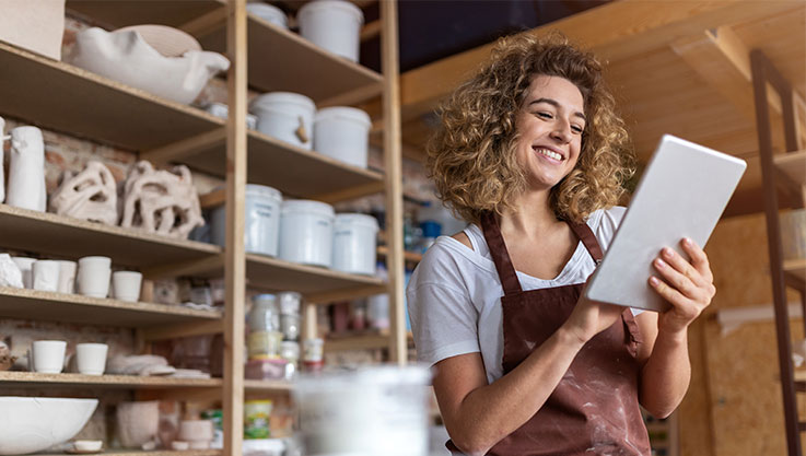 A woman stands in front of shelving filled with pottery holding a tablet.